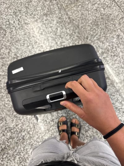 An over-the-shoulder, high-angle shot shows a person's hand gripping the telescoping handle of a black, hard-shell suitcase. The suitcase is positioned on a speckled gray floor, possibly in an airport or public transit area. The person is wearing denim shorts and black slide sandals. The background appears to be a tiled floor. The location is not specified.