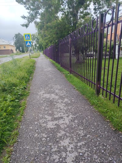 The image depicts a view of a gravel footpath running alongside a tall purple metal fence. To the left of the footpath, there is a grassy verge bordering a paved road. On the road, a single vehicle is visible in the distance. The road is lined with buildings and trees. A pedestrian crossing sign is mounted on a pole on the left side of the road. The sky is overcast. The scene is located in Bulanasch, Russia.