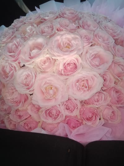 A bouquet of numerous light pink roses is presented in a close-up, top-down shot. The roses are densely packed and some appear to have small water droplets or glitter on their petals. The bouquet is wrapped in delicate pink and white fabric, creating a soft, layered effect. The background is dark and out of focus, suggesting the bouquet is being held or displayed against a dark surface. The image appears to be taken indoors with soft lighting. The image does not contain any elements that indicate a specific location in Salembaran Jati, Indonesia.