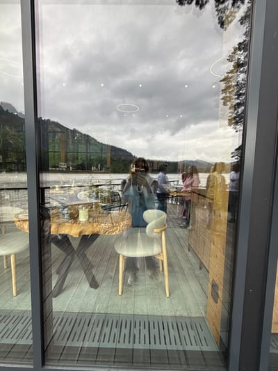 A reflective shot, likely taken from inside a building in Nizhnekayancha, Russia, looking out through a large glass window. The scene captures a natural landscape and a restaurant setting. The foreground is dominated by the reflection of an indoor dining area with a rustic wooden table, two chairs, and table settings that include a menu and a candle. Beyond this, the glass reflects an outdoor deck area with several people standing and interacting. The backdrop is a body of water, possibly a river or lake, with lush green hills and trees on the opposite bank. The sky is overcast, suggesting a cloudy day. The overall impression is one of a serene, natural setting combined with a comfortable, modern dining experience.