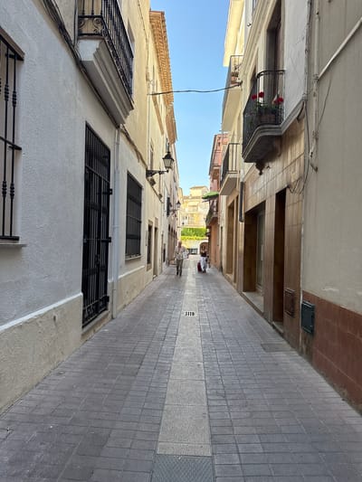 The image captures a narrow, European-style street in Tossa De Mar, Spain. The perspective is low, looking up and down the street, which is paved with gray square tiles. Buildings line both sides of the street, with their facades made of stucco and stone in varying shades of white and beige. Some buildings have balconies with decorative wrought-iron railings, one of which holds a pot of red flowers. Windows are visible, many with black wrought-iron grates.
Two people are visible in the distance, walking away from the camera. One person appears to be wearing a white shirt and dark pants, carrying a red bag. Another person is walking further down the street. A vintage-style street lamp is mounted on the side of one building. The sky is a clear, bright blue, suggesting a sunny day. The shadows cast by the buildings indicate it is likely midday. The overall impression is one of a quiet, picturesque European town.