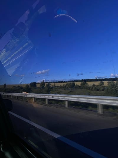 This image captures a view from inside a vehicle, looking out through the windshield and side window. The dominant subject is the bright blue sky, which takes up a significant portion of the frame. A faint, crescent-shaped moon is visible in the upper left quadrant of the sky. There are some water spots or smudges on the glass, obscuring parts of the view.

Beyond the sky, a landscape unfolds. A line of trees forms a dark green border along a field of lighter brown or yellow. A white metal guardrail runs horizontally across the mid-section of the image, separating the road from the landscape. The road itself appears dark and reflects the sky and surrounding elements. The overall scene suggests a daytime setting, likely in the late afternoon or early evening given the quality of light and the presence of the moon.

The image is taken from a moving vehicle, indicated by the slight blur of the landscape and the perspective. There are no visible people or discernible text in the image. The setting appears to be rural, with open fields and a highway or main road. The weather seems clear, with only a few small, wispy clouds visible in the sky. The mood is calm and observational, as if capturing a fleeting moment during a journey. The reflection on the car's surface adds a layer of depth and complexity to the scene.