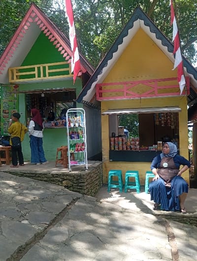 The scene depicts an outdoor commercial area, specifically in Kajar, Indonesia, featuring two small, single-story retail stalls under bright daylight.

The structure on the left has a green facade with a gabled roof accented by white and pink trim and a yellow decorative railing. An Indonesian flag, red and white striped, is attached to its roof. Inside, one individual wearing a white top and a red hijab stands facing a counter. Outside, another person in a yellow long-sleeved top and dark pants stands near a brown plastic stool.

The adjacent structure on the right has a yellow facade with a gabled roof featuring black and white trim and a pink decorative railing. An Indonesian flag is also present on its roof. A woman, wearing a dark blue hijab, a dark blue long dress, and a brown apron, is seated on a light blue plastic stool outside this stall, with her hands on her hips, looking towards the right of the frame. Two additional light blue plastic stools are positioned nearby. The stall's open front displays rows of packaged goods.

Between the two structures, on a low stone retaining wall, a white metal display rack holds various colorful packaged items. The foreground consists of an uneven, cracked paved surface composed of irregular stone or concrete slabs. Dense green foliage from trees forms the background. Shadows cast on the ground indicate a sunny day.
