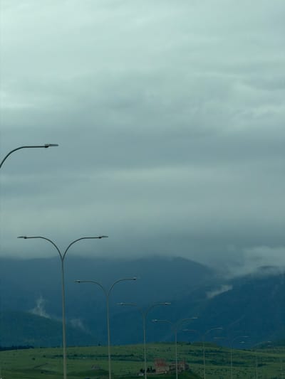 The scene is a landscape of Tedotsminda, Georgia, showing mountains, green land, and a cloudy sky. A row of curved street lights extends along the roadside.