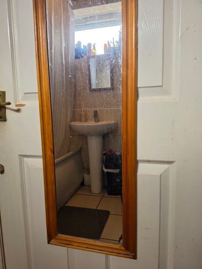 The scene, located in Shotton, United Kingdom, displays a vertical, rectangular mirror with a light brown wooden frame mounted on a white, paneled door. The door exhibits wear, including chipped paint around a brass-colored handle on the left side, and a small, round, brass-colored knob further down. The surface of the mirror is smudged or dusty in several areas.
The mirror reflects a view of a bathroom. Directly opposite the mirror, a white ceramic pedestal sink is visible, featuring a single-lever faucet. To the left of the sink, a striped shower curtain, primarily white with vertical darker lines, is partially drawn, revealing a portion of a white bathtub behind it. Above the sink, a smaller, rectangular mirror with a dark frame is affixed to the tiled wall. Above this smaller mirror, various toiletries, including bottles and brushes, are positioned on what appears to be a window sill or shelf, silhouetted against a brightly lit window. The walls around the sink are tiled in a light brown or beige color. To the right of the sink, a black waste bin with a white plastic liner is situated on the floor. The bathroom floor consists of light brown and beige tiles, upon which a dark brown bath mat is placed. No individuals are present in the image.