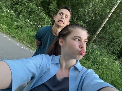 The media file is a slightly blurry, rotating, selfie-style image of two people outdoors, in what appears to be a park or natural area. The foreground shows the faces of a young woman and a young man, both looking directly at the camera. The woman, on the left, is making a kissing face, with her lips pursed and her eyes looking slightly to the side. She is wearing a light blue, textured blouse over a dark grey top. The man, on the right, has a wide-eyed, surprised expression and is grinning, showing his teeth, some of which appear to have braces. He is wearing a dark teal t-shirt.

Behind the subjects, lush green foliage and some white wildflowers are visible, suggesting a warm, possibly spring or summer day. To the right of the man, a grey paved surface, possibly a path or road, is visible. A slender tree trunk with light-colored bark is also present in the background, to the left of the woman. The lighting suggests it is daytime, likely late afternoon or early evening given the softness of the light. The overall mood of the image appears playful and informal. No text is visible in the image. The location context provided is Шевлягино, Russia.