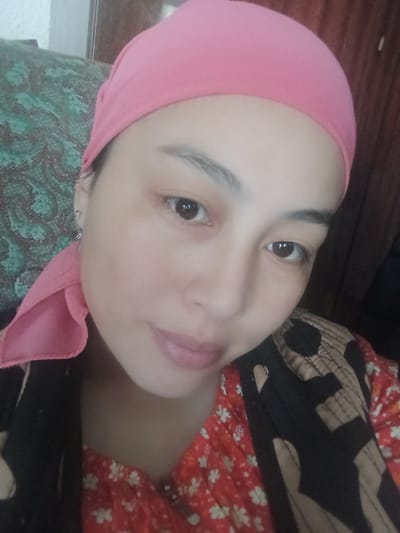 A woman is looking at the camera in a close-up shot. She is wearing a pink headscarf and a red floral shirt. She has on a patterned vest or jacket over her shirt. Her left ear is adorned with a small hoop earring. The background shows a green patterned cushion and a wooden surface. The lighting is dim, and the image quality is grainy. There is no indication of the city of Köpürö-Bazar, Kyrgyzstan.