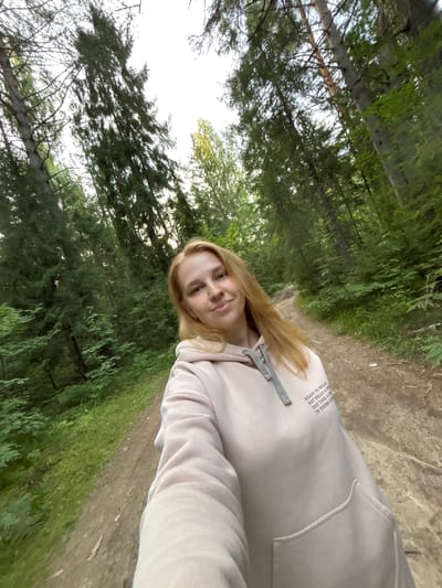A young woman with long blonde hair is taking a selfie on a dirt path in a forest. She is wearing a light pink hooded sweatshirt with gray drawstring ties and some text on the left chest. The forest is dense with tall green trees, and the path is surrounded by foliage. The sky is bright and appears overcast or hazy. The scene is located in the city of Kostiurino, Russia.