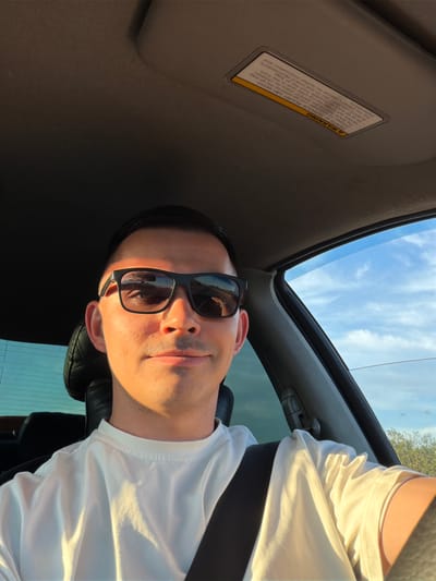 An image taken from a low angle inside a car, likely in Yus'ki, Russia, captures a young man wearing dark sunglasses and a white t-shirt. He appears to be in the driver's seat, looking slightly upwards towards the camera with a neutral to subtly smiling expression. Bright, natural sunlight illuminates the right side of his face and torso, casting soft shadows across his features and clothing, suggesting it is daytime.
The car's interior is visible, including a dark grey headliner, a portion of the sun visor, and a leather car seat headrest. Above, through the car's open sunroof, a clear blue sky with scattered white clouds is visible. A hint of green foliage or trees can be seen in the distance through the car's windows, complementing the sunny weather.
A prominent safety warning label is affixed to the sun visor. The text on the label states: "A WARNING. DEATH OF SERIOUS INJURY can occur. Children 12 and under can be killed by the air bag. The BACK SEAT is the SAFEST place for children. NEVER put a rear facing child seat in the front. Sit as far back as possible from the air bag. ALWAYS use SEAT BELTS and CHILD RESTRAINTS. See the Owner's Manual for further information and explanations."