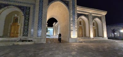 The image captures a night view of an architectural site in Satkak, Uzbekistan. The scene features a large, arched entryway made of light-colored brick, adorned with intricate blue and green mosaic tilework. To the left, a wooden door is visible within an arched alcove, also decorated with mosaic tiles. To the right, a series of smaller arched openings, likely entrances, are illuminated by warm lights, highlighting similar decorative tilework. In the foreground, a paved courtyard area is visible, with a low planter containing white flowers to the left. In the mid-ground, a person wearing dark clothing and a baby carrier on their front stands near the center of the courtyard, looking towards the right side of the scene, with their arm raised as if pointing or gesturing. Further in the background, through the large archway, another section of the complex is visible, featuring white walls and columns. The overall atmosphere is one of illuminated historical architecture at night.