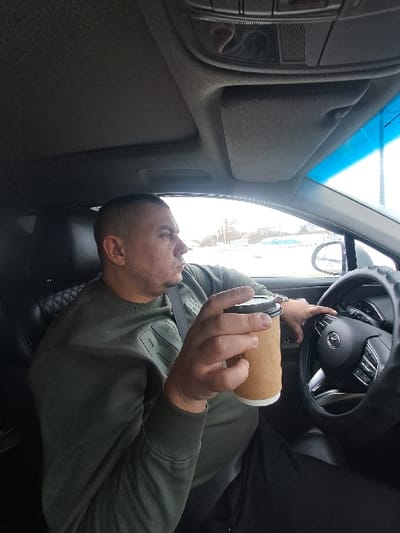 An adult male with a short haircut and stubble is seated in the driver's seat of a vehicle, looking forward and slightly to the right. He wears a dark olive green long-sleeved sweatshirt with subtle lettering and dark trousers, secured by a visible seatbelt across his chest. His right hand holds a light brown paper coffee cup with a dark lid and a white insulating sleeve. His left hand is positioned on the black steering wheel, which features a padded cover and a central Mazda logo. The car's interior includes dark seat upholstery and an overhead console with controls. Through the windshield, which has a blue tint strip at the top, and the driver's side window, a snow-covered outdoor environment is visible. This external scene features snow-laden ground, distant trees, and indistinct structures suggesting a roadside or commercial area under an overcast or bright sky. The specific location within the city of Putyatino, Russia, cannot be determined from the visual information provided in the image.