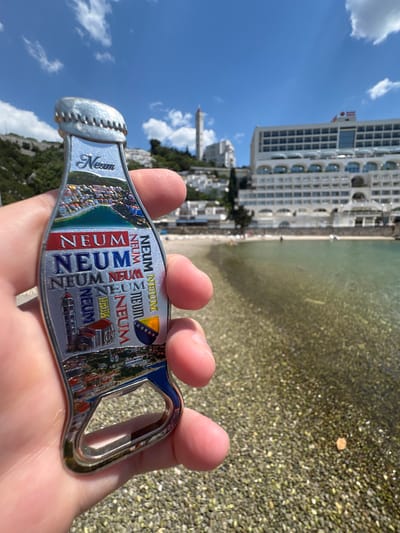 Here's a forensic description of the image:

A hand holds up a souvenir bottle opener against a sunny beach scene. The bottle opener is shaped like a bottle and features the name "Neum" in various styles and colors, along with depictions of local landmarks and the flag of Bosnia and Herzegovina.

Behind the hand and the bottle opener, the scene includes a beach with clear water, a pebbled shoreline, and a large white hotel building in the background. The hotel is multi-storied and has a modern architectural design. A hill with some trees is visible to the left of the frame. The sky is blue with scattered clouds. The scene is likely taken in Neum, Bosnia and Herzegovina.