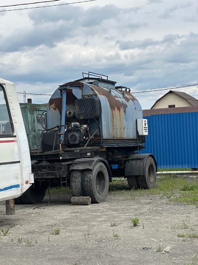 An old, rusty blue tanker truck is parked on a gravel lot next to a white vehicle, likely a bus. The tanker truck has a large cylindrical tank, a visible engine, and multiple tires. In the background, a blue corrugated metal fence and a house with a brown roof are visible. The sky is overcast with gray clouds. This scene is located in Slyudyanka, Russia.