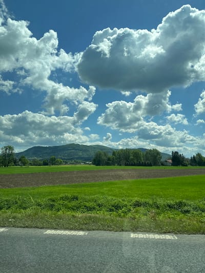 The image captures a landscape scene in or near Ustroń, Poland. The sky occupies a significant portion of the frame, filled with cumulus clouds against a blue sky. The terrain includes a field with areas of green grass and cultivated soil, bordered by trees. In the background, the slopes of a mountain are visible. A road runs along the right edge of the frame, marked with white lines. No people are visible in the scene.