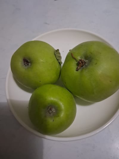 Three green apples are arranged on a white plate. The apples are round and have a slightly textured skin. One apple is positioned at the top of the plate, while the other two are arranged below it, forming a triangular shape. The background is a light-colored, textured surface, possibly a table. The lighting suggests an indoor setting, possibly in the morning or afternoon. There are no people visible in the image. The scene appears to be a simple still life.