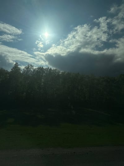 The image shows a bright, sunny sky with scattered white clouds. The sun is visible and creating a glare. A dark line of trees runs along the middle and right side of the image, obscuring much of the ground. The right edge of the image appears to be a paved surface. Location is purported to be Bezmenovo, Russia.