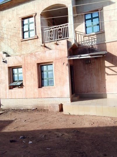The image shows the facade of a two-story building with an unfinished, textured peach-colored exterior. The building has multiple windows with green frames. On the first floor, there are two windows; one is partially open, and the other is closed. The second floor features a balcony with a white wrought-iron railing and two windows, one above and one to the right of the balcony. A simple, white canopy extends over the entrance on the first floor. The ground in front of the building is a dirt lot with scattered debris, suggesting an unpaved or under-construction environment. The lower section of the building's exterior wall has a patterned tiling. A small plant is growing near the tiled steps leading to the entrance. The lighting suggests it is daytime, with shadows cast by the building and possibly nearby structures. The scene is located in Tuntube-Tsefe, Nigeria, a rural or semi-rural area. There are no people visible in the image.