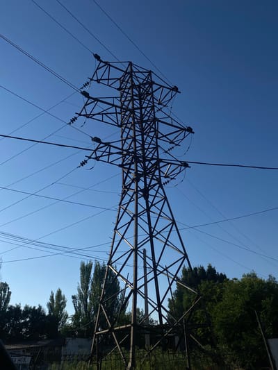 The image shows a large, lattice-like metal power line tower silhouetted against a clear blue sky. Several thick cables are suspended from the tower, crisscrossing the frame. To the right of the tower, lush green trees are visible, with their foliage extending upwards and obscuring some of the sky. The sun appears to be high in the sky, suggesting it is daytime, likely a clear and sunny day. The scene is industrial yet natural, with the man-made structure of the power lines contrasting with the organic shapes of the trees. There are no people visible in the image. Some text is visible on a sign to the far right, but it is not clearly legible. The orientation of the image suggests it was taken looking upwards towards the sky and the top of the tower. The location is specified as Төмөнкү Ала-Арча, Kyrgyzstan.