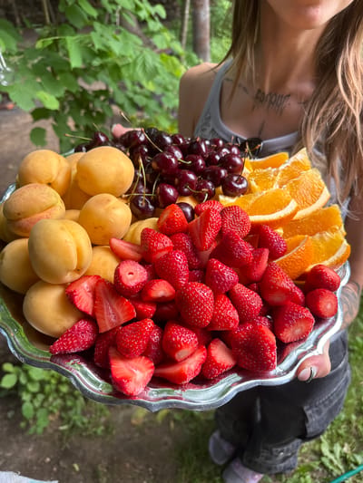 In a garden setting in Буран Снт, Russia, a person is holding a silver platter filled with a variety of fresh fruits. The platter contains whole apricots, dark cherries, sliced strawberries, and orange wedges. The person, wearing a gray tank top and dark pants, is partially visible from the chest down, with tattoos on their neck and chest. The background shows greenery, suggesting an outdoor, natural environment.