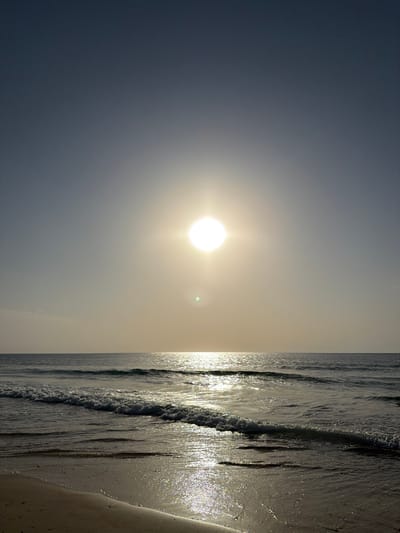 The image depicts a sunset or sunrise over the sea at Beit Yanai, Israel. The upper half of the frame shows a clear sky, transitioning from a dark blue-grey at the top to a lighter, hazy pale yellow-grey near the horizon. A brilliant, white circular sun, emitting distinct rays, is centrally positioned in the upper half of the image, with a faint green lens flare visible beneath it. The middle section of the image features the open sea with visible wave patterns. A wide, bright path of intense sunlight reflects on the water surface, extending from the horizon towards the viewer. Multiple waves are seen breaking near the shore, exhibiting white foam. The lower portion of the frame shows the wet sandy beach, also reflecting sunlight. There are no individuals, structures, or discernible objects present on the beach or in the water.