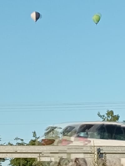 The image shows a clear blue sky with two colorful objects that appear to be hot air balloons. The balloon on the left is a translucent light green, and the balloon on the right is a bicolor design of dark gray and white. Both balloons are depicted in flight.
To the right of the balloons, a section of a white vehicle, possibly a bus or RV, is visible. The vehicle's windows are tinted and reflect the surrounding environment. The side of the vehicle has a vertical metallic structure with horizontal lines, suggesting it might be a railing or a part of the vehicle's body. Beyond the vehicle, a line of power lines stretches across the frame, and trees with green foliage can be seen. The overall lighting suggests it is daytime, likely under sunny weather conditions.
The scene is reminiscent of a bright, clear day. There are no people clearly visible. The reflection on the vehicle suggests it is stationary or moving slowly. The image's orientation is slightly tilted. The context provided places the scene in Wilkasy, Poland. No text is visible in the image.