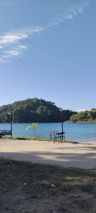 This is a landscape photograph taken on a sunny day in Calang, Indonesia. The scene depicts a tranquil waterfront with a clear blue sky and a few wispy clouds. In the foreground, a dirt path is visible, transitioning into a grassy area with scattered leaves and debris. Further back, a sandy beach meets the turquoise water of a bay.
Dominating the background is a lush, green, tree-covered hill. The calm water of the bay is the central element, reflecting the bright sky. Along the edge of the water, there are several outdoor seating arrangements. On the left, a black metal bench and a small palm tree are positioned. To the right of the palm tree, a table with four chairs, two of which are black and two are dark, is set up. Another table with chairs is visible further to the right. Two lampposts with green light fixtures stand along the waterfront, casting shadows on the ground, suggesting the time of day is likely late morning or early afternoon.
The overall mood of the image is peaceful and serene, evoking a sense of calm and relaxation. The clear weather and bright sunlight contribute to the idyllic atmosphere. There is no visible text in the image.