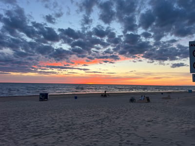 This image captures a beach scene at sunset in Narva-Jõesuu linn, Estonia. The sky is filled with dramatic clouds, transitioning from a deep blue at the top to vibrant oranges and pinks near the horizon where the sun is setting over the sea. The water is calm, with gentle waves rolling towards the sandy shore. On the beach, the sand is rippled, showing the texture left by the wind and tides.
In the middle ground, there are a few scattered objects and people. A lone figure stands near the water's edge. Further back, a bench with two bicycles parked nearby is occupied by two individuals sitting down. Other park benches and a blue trash receptacle are visible on the beach, suggesting a public recreational area. A signpost with some markings is partially visible on the right side of the frame. The overall atmosphere is peaceful and serene, characteristic of a late evening on the coast.