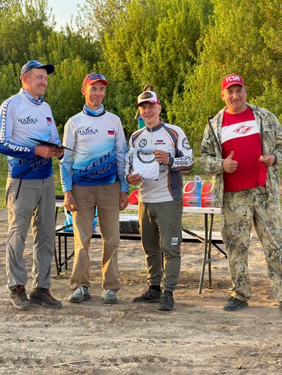 The image shows a group of four men standing outdoors on a dirt ground, possibly at a competition event. In the background, there's lush green foliage.
From top to bottom:
* The top man is wearing a red baseball cap with the letters "FCSM" and a red t-shirt with the logo of the FC Spartak Moscow soccer club, camouflage jacket, and giving a thumbs-up.
* The second man wears a white cap and white jacket, and is holding a white shirt. On a table behind, there are award trophies or plaques that read "ЧАЙКА В КОСМОСЕ", which translates to "Seagull in Space", and "1 MECT" and "2 MECTO", indicating 1st and 2nd places. The dates May 16, 2026, and May 16, 2020, are visible.
* The third man is wearing a blue and white jacket with the word "ЧАЙКА" and "FEEDER TEAM" on it. He wears a red cap.
* The bottom man is wearing a jacket with the word "ЧАЙКА" and "FEEDER TEAM" on it.
The awards indicate a connection to Krasnyi Bor and the "Seagull in Space" competition.