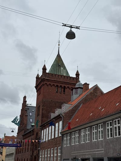 The image shows a view of a street in Elsinore, Denmark, with historic brick buildings. In the foreground, a dark gray building with white-framed windows lines the street. Above this building, a red-tiled roof is visible. To the left, a tall brick building with a green, pointed roof and spires dominates the scene. Several strings of colorful flags and banners are strung across the street. Some silhouetted figures are hanging from wires, including a green figure and a blue figure that appears to be an angel. Overhead, power lines and a street lamp are visible against a cloudy sky. The overall atmosphere is overcast.