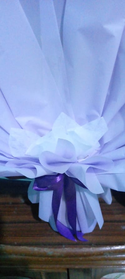 A bouquet of light purple and white tissue paper is tied with a dark purple ribbon. The bouquet is placed on a wooden surface that appears to be a table or counter. The background is blurred and a light purple color, suggesting it is also made of tissue paper or fabric. The scene is indoors, with soft lighting. No people are visible in the image. The location in the city of Uleemeuria, Indonesia is not determinable from the image itself.