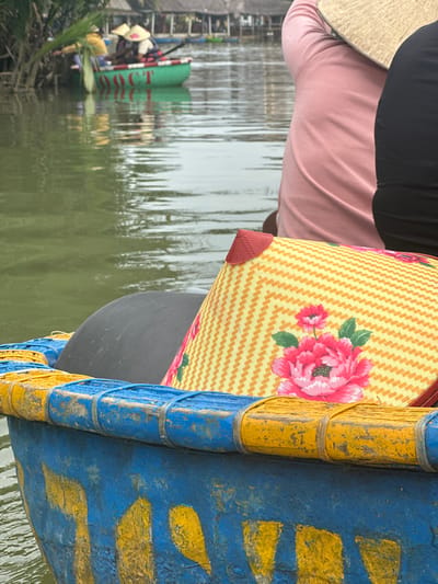 A brightly colored woven bag with a pink floral print is positioned near a blue and yellow painted boat edge. Someone wearing a traditional conical hat, a pink shirt, and black pants is partially visible behind the bag. In the background, the water reflects other boats and people. The overall scene suggests a boat ride through a waterway.