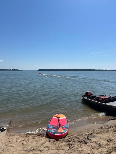 An overhead shot captures a bright, clear day on a wide, calm body of water, likely a lake or river, under a vast, unblemished blue sky. In the foreground, a sandy beach slopes gently towards the water. Resting on the sand, near the water's edge, is a red and white paddleboard with an orange paddle. To the right, a small, dark-colored motorboat with a red life jacket visible in its stern is partially submerged in the shallow water. The boat's registration number, "AB2301RUS54," is visible on its side. Further out on the water, a jet ski, with two individuals wearing orange and blue attire, creates a wake as it speeds across the surface. In the distance, the horizon is marked by a dense line of green trees, suggesting a forested shoreline. The overall scene conveys a peaceful, recreational atmosphere on a warm summer day.
