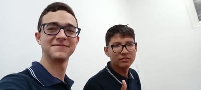 The image shows two young men standing in front of a white wall. The man on the left is wearing a dark blue polo shirt with a white stripe on the collar. He is wearing rectangular black framed glasses and is looking directly at the camera with a slight smile. The man on the right is wearing a dark blue polo shirt with a white stripe on the collar. He is wearing rectangular black framed glasses and is looking directly at the camera with a neutral expression. He is giving a thumbs up with his right hand. There is a window in the background.  The image appears to have been taken in Encarnación, Paraguay, based on the provided coordinates.