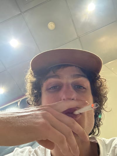 A young individual with curly brown hair, wearing a brown baseball cap and a white t-shirt, is shown from a low-angle perspective, looking directly at the camera. The person holds a lit cigarette, which has an orange glowing tip, between their fingers and to their mouth. The background consists of a white dropped ceiling with square tiles, multiple recessed bright light fixtures causing lens flare, and a single circular smoke detector. The scene is illuminated by overhead artificial light. The location is specified as Kasarvarne, India.