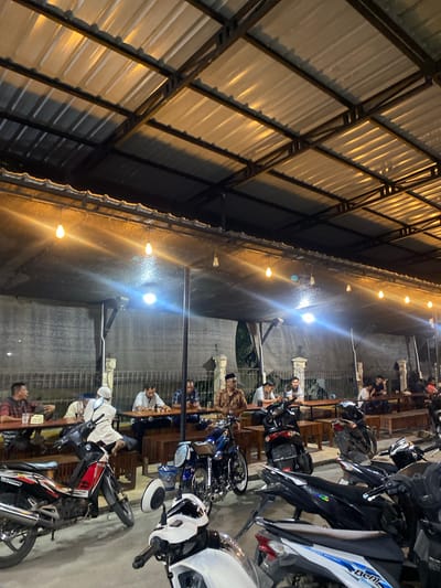 In Keudematangpanyang, Indonesia, an outdoor cafe is captured at night, illuminated by overhead lights. Several men are seated at tables, some appearing to be engaged in conversation, while others are looking down at their food or drinks. In the foreground, multiple motorcycles are parked, some with their lights on, suggesting they belong to the patrons. The cafe features a corrugated metal roof supported by a dark metal frame, with dark netting or fabric creating a partial wall on one side. The overall atmosphere is casual and social, with the presence of both diners and their vehicles indicating a typical evening at a local establishment.