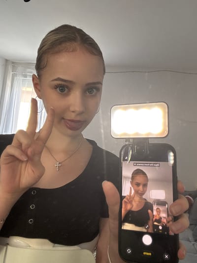 A young woman with light hair, blue eyes, and defined eyelashes is captured in a selfie. The image is taken indoors, likely in a room with light-colored walls and a window with white blinds visible in the background. The woman is wearing a black top with silver eyelet details and a silver necklace with a cross pendant. Her hair is slicked back, featuring stylized baby hairs around her temples.

She is holding a black smartphone in her left hand, which has a small, rectangular LED ring light attached to its top, casting a bright glow. With her right hand, she makes a peace sign, and her tongue is slightly sticking out, giving her a playful expression.

The smartphone's screen is visible, displaying the live preview of the selfie she is currently taking. This creates a recursive or "Droste effect" as her image, complete with the peace sign and the phone itself, is visible within the screen. On the phone screen, mirrored and rotated text reads "Clean the front camera. X" and "PHOTO." The primary light source appears to be the attached ring light, illuminating her face and the immediate surroundings. The original image is rotated 90 degrees counter-clockwise.