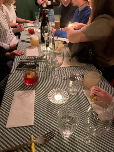 A group of at least six individuals is seated around a table draped with a green and white checkered tablecloth, in an indoor restaurant or bar setting identified as Chaîne d'Ancre in Ixelles - Elsene, Belgium. The environment is dimly lit with warm, ambient lighting. In the center-right of the frame, a person with long brown hair, wearing a light brown top, is holding a black lighter and actively igniting a small white tea light candle housed in a clear, fluted glass holder. A dark shoulder bag rests on the table beside them. To their left, another individual in a blue sweater is partially visible. On the left side of the table, a person in a white and blue striped shirt has their hands resting on the tablecloth.

The table is laden with various objects: multiple beverages including a red cocktail with a lemon twist and ice in a faceted glass, an amber-colored cocktail with foam, clear water in a bottle, and several wine glasses, some empty, others containing clear or light-colored drinks. A beer glass is visible in the background. Two dark wine bottles are present, one being handled by a person in the upper background. Cutlery, including a fork resting on a yellow chopstick holder, chopsticks, and a knife, is arranged on the table. Several white folded napkins and paper menus are also visible. A dark-colored mobile phone rests on the table on the left side. In the bottom right corner, a hand adorned with a large gold flower-shaped ring on the index finger and a silver ring on the ring finger rests near a drink.