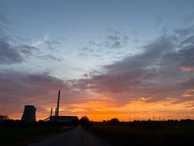 The image depicts a sunset view on a road leading towards the Bobov Dol thermal power plant in Mednikarovo, Bulgaria. The sky is a gradient of blues and oranges, with clouds dispersed throughout. The silhouette of the power plant, including cooling towers and smokestacks, is visible against the bright sky. An array of electrical power pylons are also visible in the distance. The foreground shows a paved road flanked by vegetation. The overall lighting suggests either early morning or late evening. No people are visible in the scene.