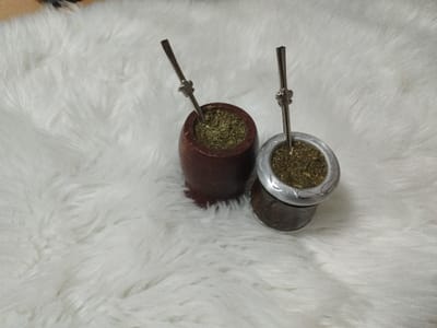 The image captures a still life scene, stated to be in al Burj, Palestinian Territory, though no specific geographical identifiers are visible within the frame. Two mate gourds, prepared with yerba mate and bombillas, are positioned on a white, fluffy faux fur surface. The gourd on the left is dark reddish-brown, appearing to be made of wood, and has a smooth, cylindrical body. The gourd on the right consists of a dark, possibly natural gourd base, topped with a light silver-colored metal rim adorned with etched or embossed decorative patterns. Both gourds are filled with light green, granular yerba mate and each contains a silver-colored metal bombilla. The bombilla in the wooden gourd has a simpler design, while the one in the silver-rimmed gourd features a small, intricate floral or star-shaped embellishment near its top. No individuals or specific actions are depicted.
