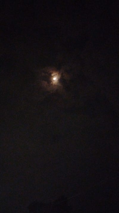 The image captures a dimly lit night sky over Banjarsari, Indonesia, with the full moon prominently visible amidst scattered clouds. The moon appears as a bright, luminous orb, partially obscured by wisps of dark, textured clouds that lend a sense of depth to the scene. The overall atmosphere is somber and tranquil, suggesting a peaceful night. The dark sky, punctuated by the moon's glow, creates a dramatic contrast. No people or specific objects are identifiable, and there is no visible text. The setting is a natural, outdoor environment at night, likely rural or suburban given the lack of urban light pollution. The weather appears to be partly cloudy, with the clouds adding a subtle, dynamic element to the celestial display.