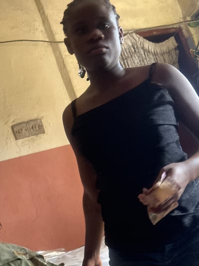 An individual stands indoors within a room in Iriebe, Nigeria. The person, with dark skin and dark braided hair, wears a black ribbed one-shoulder or spaghetti-strap top and dark trousers. A pair of dark, dangling earrings adorns the visible left ear. The subject holds a folded wad of light-colored currency notes in their right hand, pressed against their torso, and looks directly towards the camera with a neutral expression.

The room features a wall painted in two horizontal sections: a light yellow/cream upper portion and a terracotta-colored lower portion. An aged, rectangular dual electrical outlet is embedded in the upper wall section, left of the individual. In the upper right background, a light-colored, possibly patterned curtain is visible. The bottom left foreground shows draped material, potentially bedding or fabric covering an object.