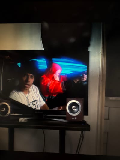 A person is depicted on a screen, with blue drapery behind them. A second person, wearing orange, is above the first person, suspended in the air. Speakers are positioned to the right of the screen. The entire scene is framed as if viewed from a monitor or television screen. There is an undefined element on the left side, partially obscuring the screen's display.