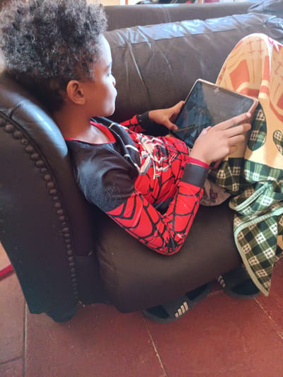 The image shows a young person with dark, curly hair lying on a dark leather couch. They are wearing a red and black long-sleeved shirt with a spiderweb pattern, likely a Spider-Man costume. The person is holding a tablet computer in their hands and appears to be looking at it. They are covered by a light-colored blanket with an orange and white geometric pattern and a green and white plaid blanket. The couch has decorative nailhead trim along its edge. The floor in front of the couch is reddish-brown and appears to be tiled or made of stone. There are also black slide sandals visible on the floor near the person's feet. The setting appears to be a living room. Based on the provided image information, the location is in Saurimo, Angola.