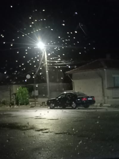 The image captures a night scene in the city of Gradina, Bulgaria, likely a residential or suburban area. A black car is parked on a wet street illuminated by a bright street lamp. The street appears to be asphalt and has puddles, suggesting recent rain. In the background, a single-story building with a sloped roof is visible, its facade a light color. To the left of the car, a bush or small tree with green foliage is present. The sky is dark, with numerous circular light refractions, indicative of either raindrops or dust particles on the camera lens, superimposed over the scene, particularly near the street lamp. The overall impression is one of a quiet, possibly damp, evening in a Bulgarian town.