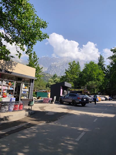 This image depicts a street scene in Arstanbap, Kyrgyzstan, with a clear blue sky and fluffy white clouds dominating the upper portion. In the foreground, a paved road with white lane markings is visible, partially shaded by the leaves of trees. To the left, a small, modern-looking shop with a glass storefront displays various items, including what appear to be toys and household goods. Adjacent to the shop, a black building with a dark roof stands. Further down the road, several cars are parked, including a dark gray SUV in the foreground. A person in a light blue shirt is walking along the sidewalk near the parked cars. In the background, lush green trees line the street, leading up to a majestic, rocky mountain range under a bright sky. The overall scene suggests a sunny day in a town nestled against a mountainous backdrop.
