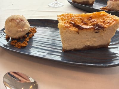 The image displays a dessert setting within what appears to be a dining establishment in Colunga, Spain. The primary focus is on a dark blue, oblong ceramic plate presenting two distinct desserts. On the right, a substantial slice of cheesecake is featured, characterized by a creamy, light-colored body and a deeply caramelized, unevenly browned top crust. A darker, thinner base is visible beneath the cheesecake. On the left side of the plate, a single scoop of light-colored ice cream, possibly vanilla or cream-based with subtle reddish swirls, rests alongside scattered, coarse, reddish-brown crumbs, likely biscuit or streusel.

In the foreground, a silver spoon with a reflective surface is partially visible in the bottom-left corner. The entire scene is set on a light-colored, possibly off-white or beige, textured fabric tablecloth. In the out-of-focus background, another dark, oblong plate holds a second dessert, partially obscured, and the base and stem of a clear drinking glass are visible. No individuals are present or engaged in any actions within the frame.