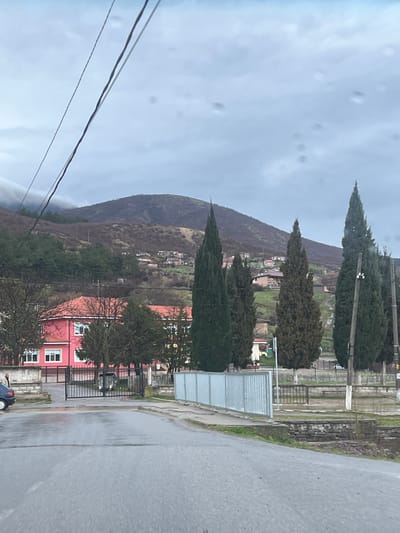 The image depicts a rural or semi-rural scene in Sotirya, Bulgaria, on an overcast day, likely observed from a vehicle on a wet asphalt road.
In the immediate foreground, the perspective is from a wet, gray asphalt road that curves slightly to the right. Overhead, multiple dark utility wires traverse the cloudy, gray sky. To the left, a portion of a dark blue vehicle, possibly a sedan, is visible. Adjacent to the vehicle, a male individual stands facing right, wearing a light-colored jacket. To the right of the road, a silver corrugated metal barrier/fence, approximately waist-high, extends from the foreground towards the midground. Beyond this fence is a low stone wall and a patch of dry ground.
The midground features a prominent two-story building with a distinctive reddish-pink exterior and white-framed rectangular windows, likely a public or institutional structure, situated behind a black metal gate. Several bare deciduous trees and dark green evergreen trees, some of which are tall and slender like cypresses, surround this building. To the right of the road, a line of these tall, narrow evergreen trees extends vertically, obscuring parts of the background. Wooden utility poles are interspersed among these trees.
The background is dominated by a large, undulating hill or mountain covered with sparse, dry vegetation and some clusters of darker evergreen trees, suggesting a late autumn or winter season. Numerous small houses and other structures with varying roof colors are scattered across the lower slopes of the hill, indicating a residential area or village settlement. The sky is uniformly overcast with a light gray tone, and several circular lens flares or smudges are visible in the upper half of the image.