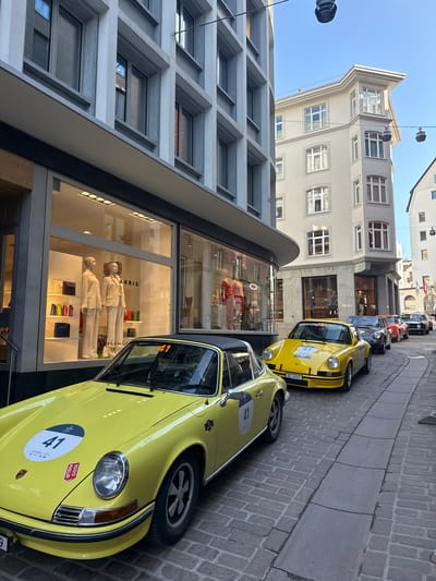 The image depicts a narrow, cobblestone street in Sankt Moritz, Switzerland, filled with vintage sports cars and flanked by luxury retail buildings under a clear blue sky.

In the foreground, a bright yellow Porsche 911 sports car is prominently parked on the right side of the street. It features a black soft-top roof. Its hood displays a white circular decal with the number "41" in black, a "CHOPARD" logo, and a red "1000 MIGLIA" sticker. The front bumper has a Swiss license plate, "SZ 4311". Immediately behind it, another bright yellow Porsche 911 is parked, bearing a white circular decal with "42" on its windshield and "SUPERCARROWNSCIRCLE.COM" text. A line of additional vintage Porsche 911s, in colors including dark grey/black, red, and dark green/black, extends down the street, all similarly marked with event decals, suggesting a rally or car show.

To the left, a modern, multi-story building with a grey concrete facade features large, illuminated display windows. One window prominently displays "AKRIS" branding and showcases two pale mannequins dressed in beige suits, surrounded by an array of colorful handbags on shelves. Further along, "HANRO" branding is visible in another window. On the right, across the street from the parked cars, stands an older, lighter-colored multi-story building with a distinctive corner architectural style and an overhanging roofline. One of its ground-floor windows displays "PRADA" branding. The street itself is paved with small, grey cobblestones and features a central, linear drainage channel. Dark, circular street light fixtures hang from overhead wires against the clear blue sky. No individuals are actively visible interacting with the vehicles or the immediate street scene.