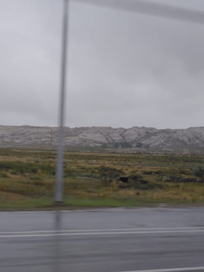 The image depicts a vast, overcast landscape with a focus on a rugged, mountainous terrain in the background. The foreground features a wet asphalt road with white lane markings, indicating recent rainfall. Beyond the road, a grassy field stretches out, dotted with some sparse vegetation and what appear to be remnants of wooden structures or fences. A tall, thin pole, possibly a utility pole, stands vertically in the middle ground, partially obscuring the view of the distant mountains. The sky is uniformly gray and cloudy, suggesting a cool or damp day. The scene appears to be set in a rural or semi-arid environment, characteristic of the steppe regions of Kazakhstan. The location is identified as within the Arshaly rural district of Kazakhstan. There are no people or animals visible in the image.
