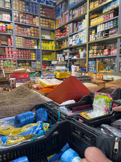 This image displays the interior of a densely packed market stall or small grocery store located in Ksar-el-Kebir, Morocco. The scene is characterized by vertical metal shelving units that line the walls, fully stocked with a diverse array of packaged consumer goods, including cereals, canned items, bottled liquids, and various boxed products.

In the foreground and mid-ground, a counter surface is covered with large, open displays of bulk items. A prominent, conical mound of deep red powder, consistent with paprika or chili powder, is centrally located. Adjacent to this, piles of other granular goods are visible, including a lighter brown, granular spice (possibly cumin seeds) and various nuts and dried goods. Specific packaged products, such as boxes labeled "Pruneaux La Famille" (prunes) and jars resembling chocolate hazelnut spread, are also present on the counter. A small, grey weighing scale is positioned behind the bulk spice displays.

The immediate foreground contains black plastic utility crates. These crates hold miscellaneous merchandise, including multiple rolls of blue plastic twine or rope, yellow plastic objects, a small brush with purple bristles, and clear plastic bags, one of which features pink cow branding and appears to contain small, individually wrapped items. A human hand, showing a thumb and forefinger, is visible on the right side of the image, positioned near a black zippered pouch or wallet, indicating a person interacting with the items in the foreground crates. The lighting in the scene is bright and artificial, illuminating the merchandise.
